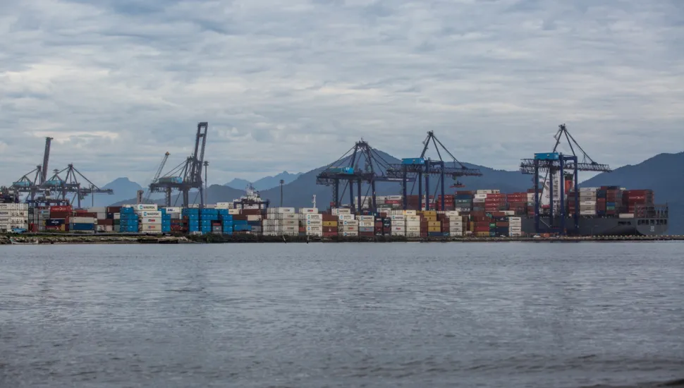 Fotografia em enquadramento amplo e vista panorâmica de um terminal portuário às margens de uma baía. Em primeiro plano, ocupa a metade inferior da imagem a superfície da água em tom cinza-azulado com pequenas ondas suaves. No plano intermediário, estende-se a faixa do porto com milhares de contêineres coloridos empilhados — predominantemente azul, vermelho, branco e amarelo — e um navio cargueiro atracado à direita. Diversos guindastes e portêineres de grande porte erguem-se ao longo da extensão do cais. Ao fundo, a paisagem é delimitada pela silhueta de montanhas sob um céu totalmente coberto por nuvens cinzentas e densas em iluminação difusa diurna.