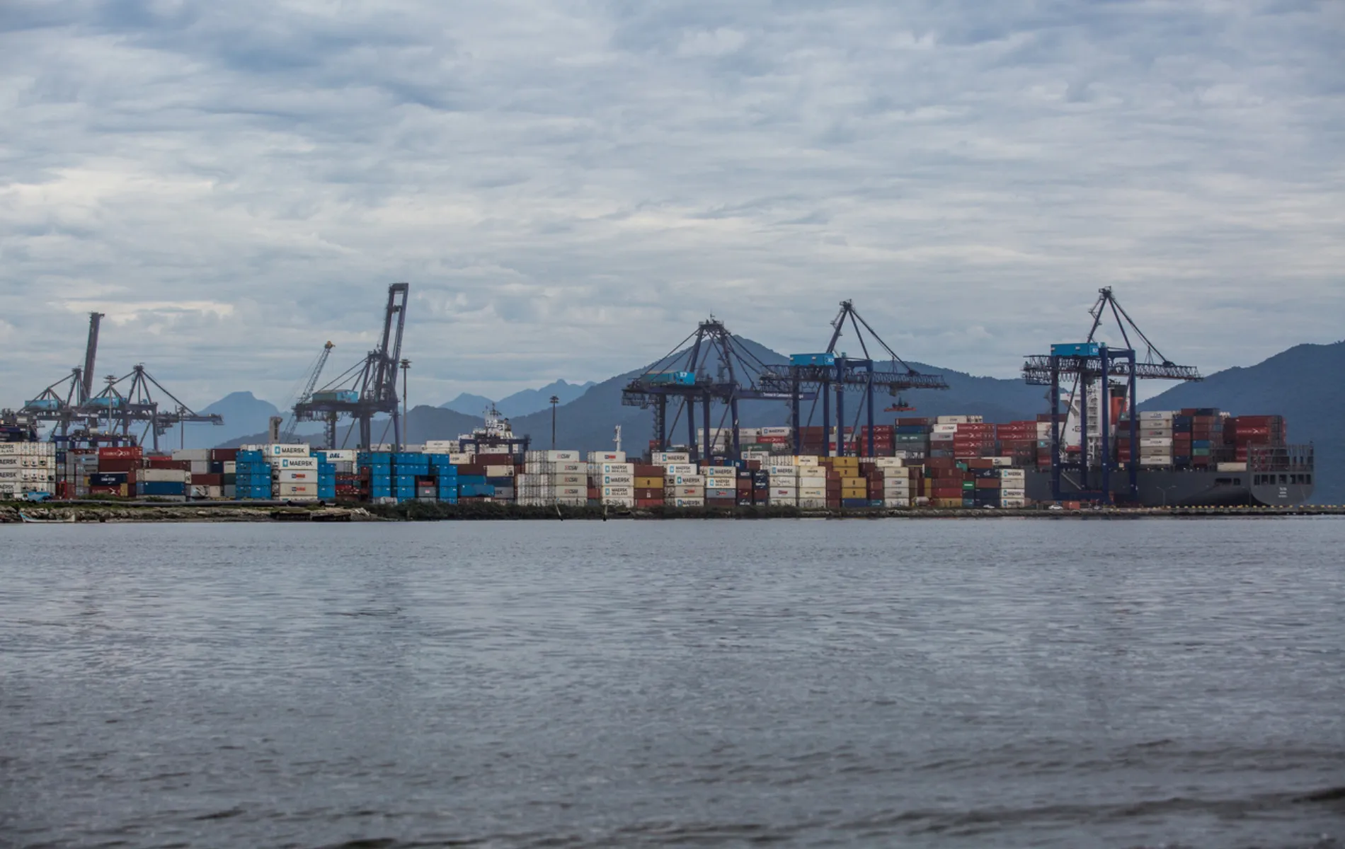 Fotografia em enquadramento amplo e vista panorâmica de um terminal portuário às margens de uma baía. Em primeiro plano, ocupa a metade inferior da imagem a superfície da água em tom cinza-azulado com pequenas ondas suaves. No plano intermediário, estende-se a faixa do porto com milhares de contêineres coloridos empilhados — predominantemente azul, vermelho, branco e amarelo — e um navio cargueiro atracado à direita. Diversos guindastes e portêineres de grande porte erguem-se ao longo da extensão do cais. Ao fundo, a paisagem é delimitada pela silhueta de montanhas sob um céu totalmente coberto por nuvens cinzentas e densas em iluminação difusa diurna.