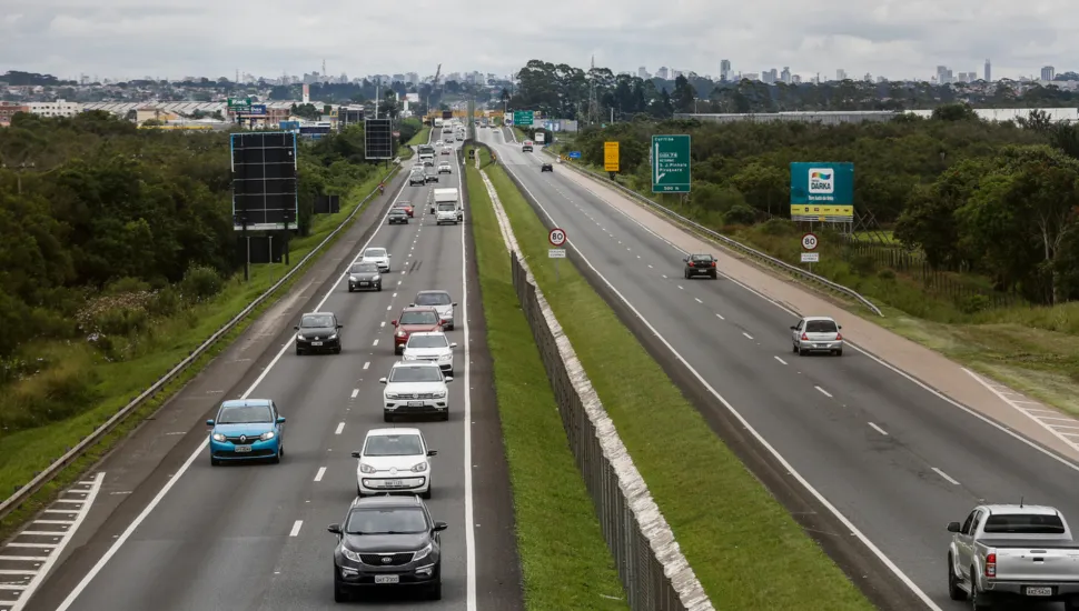 Fotografia em enquadramento amplo e plano geral aéreo de uma rodovia de pista dupla em ambiente periurbano. A via é dividida ao centro por um canteiro central gramado com mureta de proteção em concreto. À esquerda, as faixas mostram fluxo intenso de veículos — incluindo carros de passeio e utilitários — aproximando-se em direção ao primeiro plano. À direita, a pista oposta apresenta trânsito mais espaçado, com veículos afastando-se. Placas de sinalização viária com limite de velocidade de "80" km/h e painéis publicitários margeiam a rodovia sobre faixas de vegetação nativa.