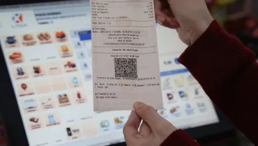 Fotografia em primeiro plano e plano detalhe das mãos de uma pessoa vestindo uma blusa de manga longa vermelha, segurando um comprovante fiscal impresso em papel térmico branco. Centralizada na imagem, a nota exibe detalhes impressos de uma compra de "GASOLINA COMUM", o valor total de 20,00 reais, a chave de acesso e um código QR Code impresso no terço inferior. Ao fundo, em profundidade de campo rasa e desfocada, vê-se a tela de um monitor de computador exibidora do sistema de ponto de venda (PDV) com diversos ícones coloridos de produtos.