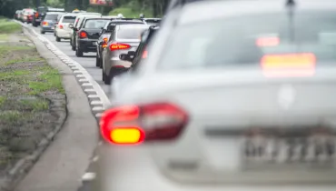 Fotografia em enquadramento médio com foco seletivo em profundidade de campo rasa, mostrando um congestionamento de veículos em uma rodovia ou via expressa. Em primeiro plano, à direita e desfocado, vê-se a traseira de um carro de passeio na cor prata, com a lanterna traseira vermelha acesa. No plano intermediário e ao fundo, em foco nítido, estende-se uma longa fila indiana de carros de diversos modelos e cores — como preto, cinza e branco — parados no tráfego. As luzes de freio vermelhas de alguns veículos estão acesas. À esquerda, a pista é delimitada por uma faixa de meio-fio zebrada em branco e cinza, margeada por vegetação rasteira e árvores ao fundo sob iluminação diurna.