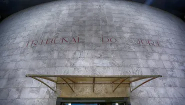 Fotografia em contra-plongée (ângulo de baixo para cima) da fachada em formato curvo de um prédio público. A estrutura é revestida por placas quadradas de mármore cinza-claro com textura marmorizada. No centro da fachada, gravada em letras caixa de tom metálico ou avermelhado, destaca-se a inscrição "TRIBUNAL DO JÚRI". Na parte inferior, encontra-se uma marquesa/cobertura de vidro sustentada por estrutura metálica sobre a porta de entrada. No reflexo do vidro da porta de acesso, veem-se a vegetação e o céu sob iluminação diurna.