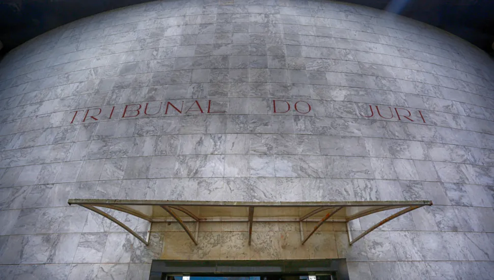 Fotografia em contra-plongée (ângulo de baixo para cima) da fachada em formato curvo de um prédio público. A estrutura é revestida por placas quadradas de mármore cinza-claro com textura marmorizada. No centro da fachada, gravada em letras caixa de tom metálico ou avermelhado, destaca-se a inscrição "TRIBUNAL DO JÚRI". Na parte inferior, encontra-se uma marquesa/cobertura de vidro sustentada por estrutura metálica sobre a porta de entrada. No reflexo do vidro da porta de acesso, veem-se a vegetação e o céu sob iluminação diurna.