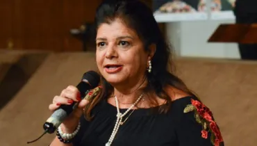Fotografia em primeiro plano da empresária Luiza Helena Trajano durante um discurso ou apresentação em um evento institucional. Centralizada no enquadramento, a mulher de pele clara, olhos castanhos e cabelos escuros na altura dos ombros segura um microfone preto próximo à boca com a mão direita, que exibe unhas pintadas de vermelho e pulseiras nos pulsos. Ela veste uma blusa preta de ombro a ombro com bordados florais vermelhos e verdes na manga e usa um colar duplo de pérolas e brincos pequenos. Ao fundo, a ambientação do recinto apresenta painéis de madeira e estruturas claras desfocadas em profundidade de campo rasa sob iluminação interna.
