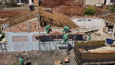 Fotografia em enquadramento amplo com plano geral elevado (plongée) de uma obra de construção e reforma residencial. Em primeiro plano, no canto inferior direito, destaca-se a caçamba de um caminhão carregado com areia e materiais de construção. Ao centro e no canto inferior esquerdo, sobre o solo de cascalho, um pedreiro de uniforme verde trabalha ao lado de um carrinho de mão e pilhas de tijolos de barro. No plano intermediário, o foco principal é a estrutura da casa em alvenaria. Três operários vestindo uniformes verdes trabalham sobre andaimes de madeira, assentando tijolos cerâmicos nas paredes externas em fase de elevação. O telhado da residência apresenta a armação de madeira exposta e sem telhas. Ao fundo, sob luz solar direta, o terreno do entorno exibe solo de terra vermelha, entulhos e outras residências vizinhas.