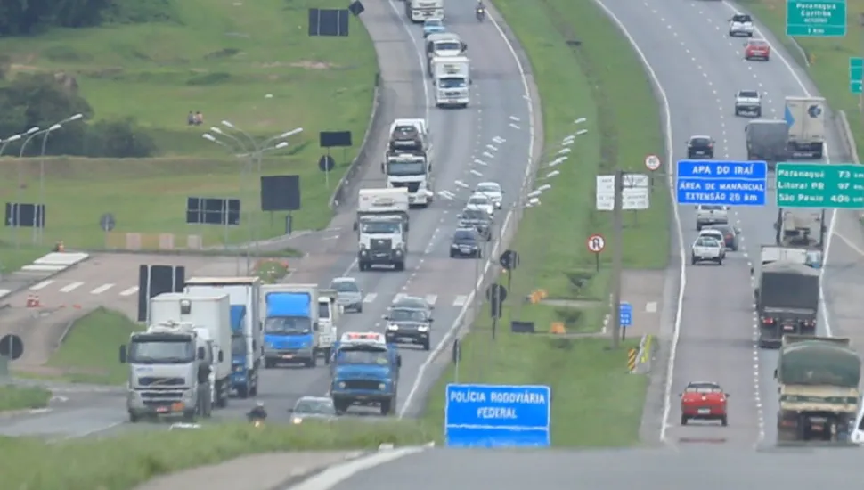 Fotografia em enquadramento amplo com plano geral aéreo de um trecho de rodovia federal de pista dupla sob iluminação diurna. Em primeiro plano, no centro, sobressai uma placa azul com a inscrição em branco "POLÍCIA RODOVIÁRIA FEDERAL". À esquerda, na pista em sentido aproximação, observa-se uma fila contínua de veículos, predominantemente caminhões de grande porte, baús e utilitários, trafegando em direção ao primeiro plano. À direita, na pista de sentido oposto, carros de passeio e caminhões seguem afastando-se. Entre as vias e nas margens, há canteiros gramados e diversas placas de sinalização verde e azul indicando rotas e trechos como "APA DO IRAÍ". Ao fundo, a rodovia estende-se em aclive suave contornada por vegetação e morros ao longe.
