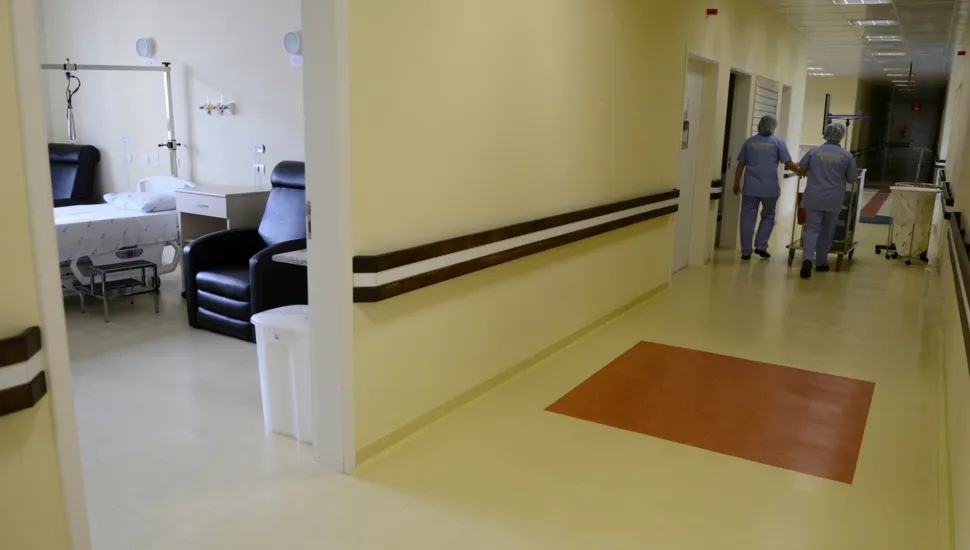 Fotografia em enquadramento amplo com plano geral de um corredor hospitalar e a vista parcial de um quarto de internação à esquerda. Em primeiro plano, à esquerda, através da porta aberta, observa-se o interior do quarto com uma cama hospitalar ajustável coberta por lençóis brancos, uma poltrona de descanso preta em couro, uma mesa de cabeceira e uma lixeira branca. No plano intermediário e ao fundo, à direita, estende-se o corredor bem iluminado com paredes amarelas claras e corrimão duplo de proteção em tom escuro. Duas profissionais de saúde vestindo pijama cirúrgico azul e touca descartável caminham de costas no corredor empurrando um carrinho de apoio. O piso claro exibe um detalhe retangular na cor terracota e a iluminação no teto é distribuída uniformemente.