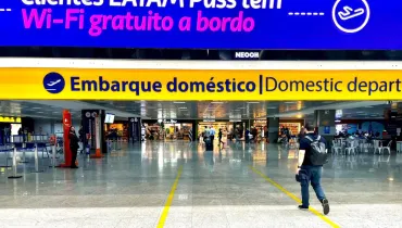 Fotografia em enquadramento amplo com plano geral do saguão de um aeroporto, destacando o acesso à área de embarque. Na parte superior, sobressai um grande painel digital azul com texto promocional em branco e rosa: "Clientes LATAM Pass têm Wi-Fi gratuito a bordo". Logo abaixo, uma faixa amarela horizontal exibe a sinalização em letras azuis e pretas: "Embarque doméstico | Domestic departures", acompanhada pelo ícone de um avião decolando. Em primeiro plano, no canto inferior direito, um homem vestindo camiseta escura, calça jeans e mochila nas costas caminha de costas em direção ao portão de acesso, sobre o piso claro e polido marcado por faixas amarelas no chão. No plano intermediário e ao fundo, observam-se tótens de atendimento, balcões de controle, passageiros circulando e lojas comerciais do terminal sob iluminação artificial interna.