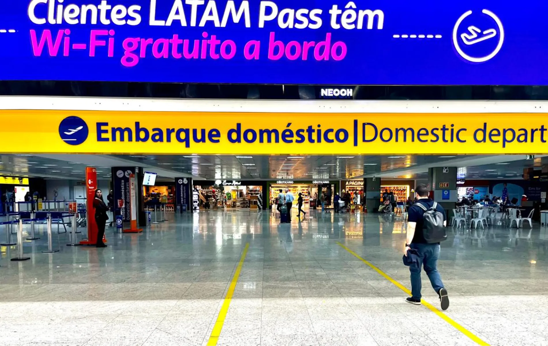 Fotografia em enquadramento amplo com plano geral do saguão de um aeroporto, destacando o acesso à área de embarque. Na parte superior, sobressai um grande painel digital azul com texto promocional em branco e rosa: "Clientes LATAM Pass têm Wi-Fi gratuito a bordo". Logo abaixo, uma faixa amarela horizontal exibe a sinalização em letras azuis e pretas: "Embarque doméstico | Domestic departures", acompanhada pelo ícone de um avião decolando. Em primeiro plano, no canto inferior direito, um homem vestindo camiseta escura, calça jeans e mochila nas costas caminha de costas em direção ao portão de acesso, sobre o piso claro e polido marcado por faixas amarelas no chão. No plano intermediário e ao fundo, observam-se tótens de atendimento, balcões de controle, passageiros circulando e lojas comerciais do terminal sob iluminação artificial interna.