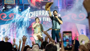 Fotografia em enquadramento amplo e plano americano da dupla sertaneja Fernando & Sorocaba apresentando-se em um show ao vivo. Em primeiro plano, a plateia aparece de costas em contra-luz, com braços levantados, chapéus de boiadeiro e um celular registrando a apresentação. No plano intermediário, sobre o palco iluminado por canhões de luz branca e azul, os dois cantores realizam o show. À esquerda, Fernando veste camiseta estampada de tom claro, segura uma guitarra elétrica bege e canta ao microfone. À direita, Sorocaba veste camisa jeans, calça escura e cinto fivelado, segurando um microfone em uma das mãos e erguendo um chapéu de boiadeiro bege com a outra mão. Ao fundo, a estrutura cenográfica do palco exibe um painel em vermelho e azul com a inscrição "CHURRASCO ON FIRE" e elementos decorativos.