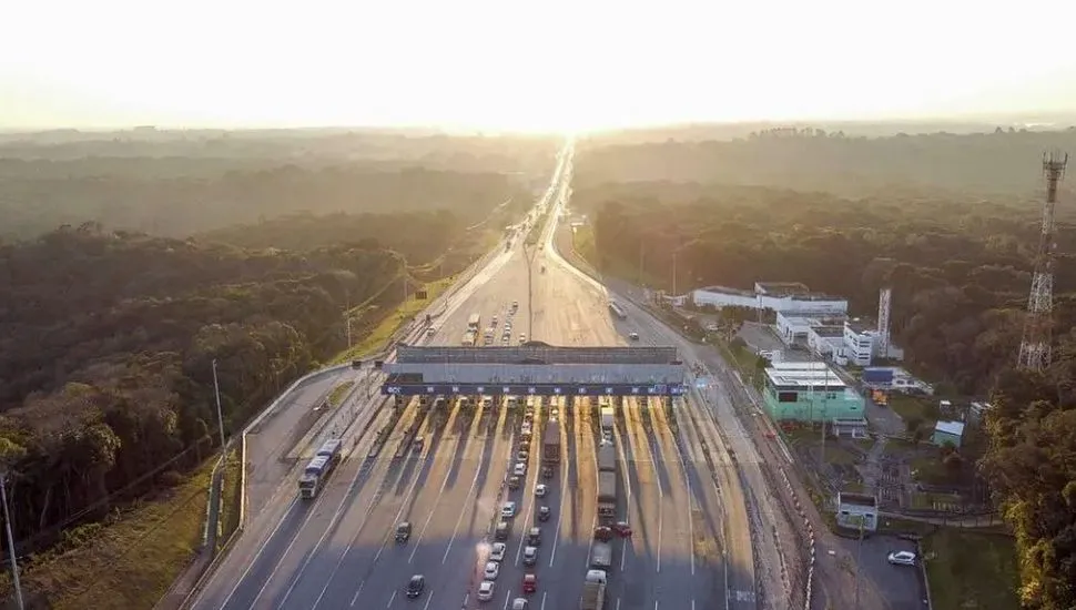 A imagem mostra uma praça de pedágio, com filas de carros e caminhões parados, com um sol de fim de tarde ao fundo e árvores ao redor.