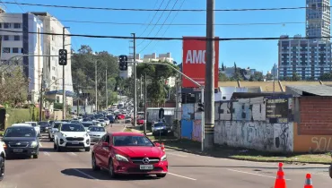 Fotografia em enquadramento amplo com plano geral de uma via urbana com tráfego intenso de veículos em um dia ensolarado. Em primeiro plano, no canto inferior direito, alinham-se cones de sinalização laranjas com faixas refletivas sobre o asfalto. Ao centro-esquerda, destaca-se um carro de passeio vermelho em movimento no cruzamento. No plano intermediário, atrás do veículo vermelho, uma fila contínua de carros brancos, cinzas e pretos estende-se ao longo da pista, aguardando o fluxo do trânsito próximo a semáforos instalados em hastes metálicas. À direita, observa-se um muro com pichações e uma placa comercial com a inscrição "SOLDA IMPERIAL", ao lado de um poste de iluminação pública e fiação aérea. Ao fundo, sob céu azul e limpo, sobressai um grande outdoor vermelho com a palavra "AMOR" em letras brancas, com vegetação de morro e edifícios altos completando o cenário urbano.