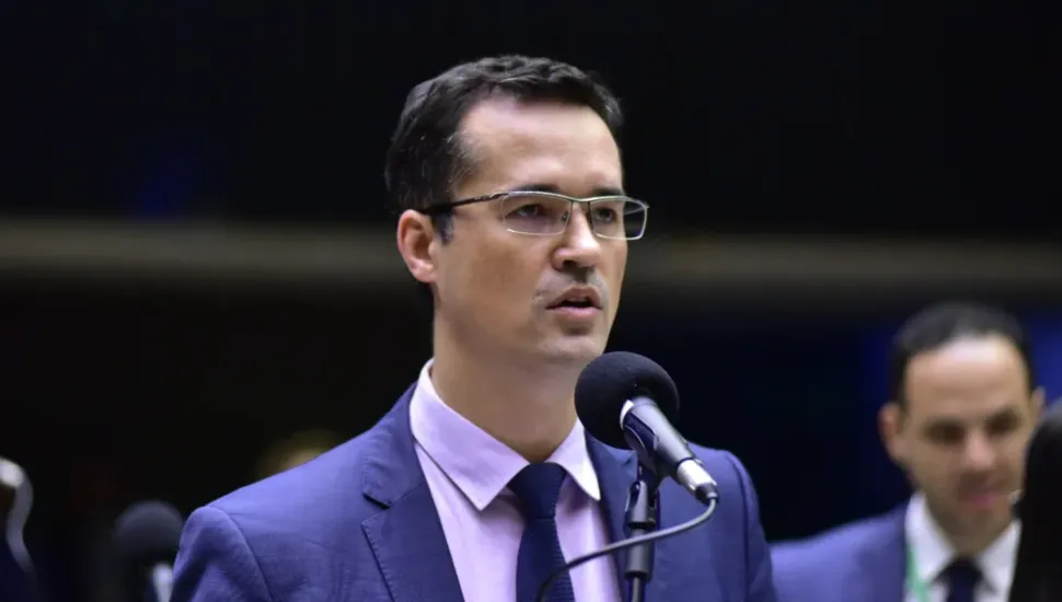 Fotografia em primeiro plano e plano médio do ex-deputado e ex-procurador Deltan Dallagnol durante pronunciamento público. Centralizado no enquadramento e levemente voltado para a direita, o homem de pele clara, cabelos curtos e escuros e óculos de grau com armação retangular fala diante de um microfone pedestal. Ele veste terno azul-marinho, camisa social lilás-claro e gravata azul-escura. Ao fundo, à direita e desfocado pela profundidade de campo rasa, observa-se outro homem de terno sob iluminação interna de plenário.