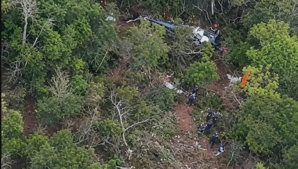 Fotografia aérea em enquadramento amplo de um local de queda de aeronave em meio a uma área de mata densa e vegetação fechada. Na parte superior direita, entre as árvores, encontram-se os destroços retorcidos da fuselagem de um helicóptero ou pequena aeronave. A clareira aberta no solo expõe a terra batida e galhos quebrados resultantes do impacto. No plano intermediário e no canto inferior direito, equipes de resgate e peritos com uniformes escuros caminham pelo terreno e realizam os trabalhos de inspeção e busca no local. Ao redor de toda a cena, árvores de copas verdes e arbustos cobrem a maior parte do terreno sob iluminação diurna natural.