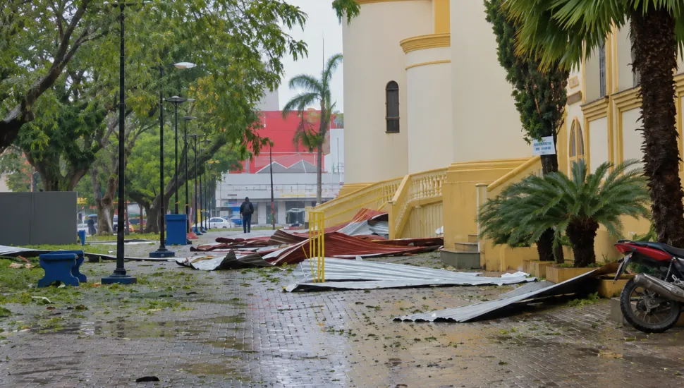 Fotografia em enquadramento amplo com plano geral do pátio externo de uma edificação histórica amarela após a passagem de uma tempestade ou vendaval. Em primeiro plano, o chão pavimentado e molhado está repleto de folhas espalhadas e folhas de telha metálica galvanizada e avermelhada contorcidas e arrancadas, caindas sobre o passeio. No canto inferior esquerdo, observa-se um banco de concreto azul e, no canto inferior direito, a roda traseira de uma motocicleta vermelha estacionada junto a palmeiras e arbustos. No plano intermediário, à direita, destaca-se a fachada de arquitetura clássica pintada em tom amarelo-claro com detalhes e escadaria lateral. À esquerda, alinham-se postes de iluminação pública pretos e árvores de copa densa ao longo do calçadão, onde uma pessoa caminha ao fundo. Ao fundo, sob céu encoberto e iluminação diurna difusa, avistam-se edifícios urbanos e vegetação.