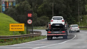 Fotografia em enquadramento amplo com plano geral do tráfego em um trecho curvo de rodovia ladeado por sinalização de trânsito. Em primeiro plano, no canto esquerdo, sobre um talude gramado, destaca-se uma placa amarela de sinalização viária com a inscrição "MEDIDOR DE VELOCIDADE" em letras pretas. Logo atrás, um radar eletrônico exibe em visor digital vermelho o número "82" e, abaixo, uma placa circular indica o limite máximo de velocidade de "60 km/h". No plano intermediário, um caminhão-guincho transporta na plataforma um carro de passeio branco, trafegando pela pista asfaltada ao lado de uma mureta metálica de proteção (guard-rail). À direita do caminhão, outro automóvel prata segue no mesmo sentido. Ao fundo, a rodovia estende-se cercada por vegetação densa e árvores sob céu encoberto e luz diurna difusa.