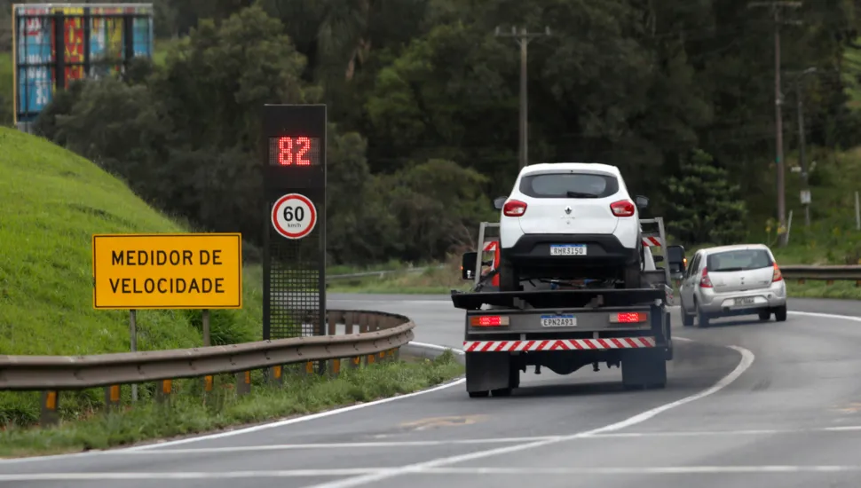 Fotografia em enquadramento amplo com plano geral do tráfego em um trecho curvo de rodovia ladeado por sinalização de trânsito. Em primeiro plano, no canto esquerdo, sobre um talude gramado, destaca-se uma placa amarela de sinalização viária com a inscrição "MEDIDOR DE VELOCIDADE" em letras pretas. Logo atrás, um radar eletrônico exibe em visor digital vermelho o número "82" e, abaixo, uma placa circular indica o limite máximo de velocidade de "60 km/h". No plano intermediário, um caminhão-guincho transporta na plataforma um carro de passeio branco, trafegando pela pista asfaltada ao lado de uma mureta metálica de proteção (guard-rail). À direita do caminhão, outro automóvel prata segue no mesmo sentido. Ao fundo, a rodovia estende-se cercada por vegetação densa e árvores sob céu encoberto e luz diurna difusa.