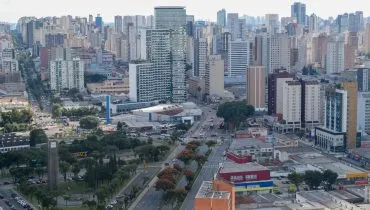 Fotografia aérea em enquadramento amplo com plano geral panorâmico da paisagem urbana de uma grande cidade sob iluminação diurna. Em primeiro plano, na parte inferior esquerda, destaca-se uma praça arborizada com canteiros verdes, vias internas de circulação e veículos estacionados. À direita, avenidas largas com múltiplas faixas cortam a área urbana, apresentando tráfego de carros e ônibus entre galpões comerciais de alvenaria e telhados claros. No plano intermediário, observa-se o cruzamento de vias principais e uma grande concentração de edifícios residenciais e comerciais de portes variados, incluindo postos de combustível e estruturas de médio porte. Ao fundo, a linha do horizonte estende-se com uma densa massa de arranha-céus e prédios altos sob céu claro e levemente ensolarado.