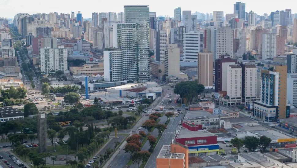 Fotografia aérea em enquadramento amplo com plano geral panorâmico da paisagem urbana de uma grande cidade sob iluminação diurna. Em primeiro plano, na parte inferior esquerda, destaca-se uma praça arborizada com canteiros verdes, vias internas de circulação e veículos estacionados. À direita, avenidas largas com múltiplas faixas cortam a área urbana, apresentando tráfego de carros e ônibus entre galpões comerciais de alvenaria e telhados claros. No plano intermediário, observa-se o cruzamento de vias principais e uma grande concentração de edifícios residenciais e comerciais de portes variados, incluindo postos de combustível e estruturas de médio porte. Ao fundo, a linha do horizonte estende-se com uma densa massa de arranha-céus e prédios altos sob céu claro e levemente ensolarado.