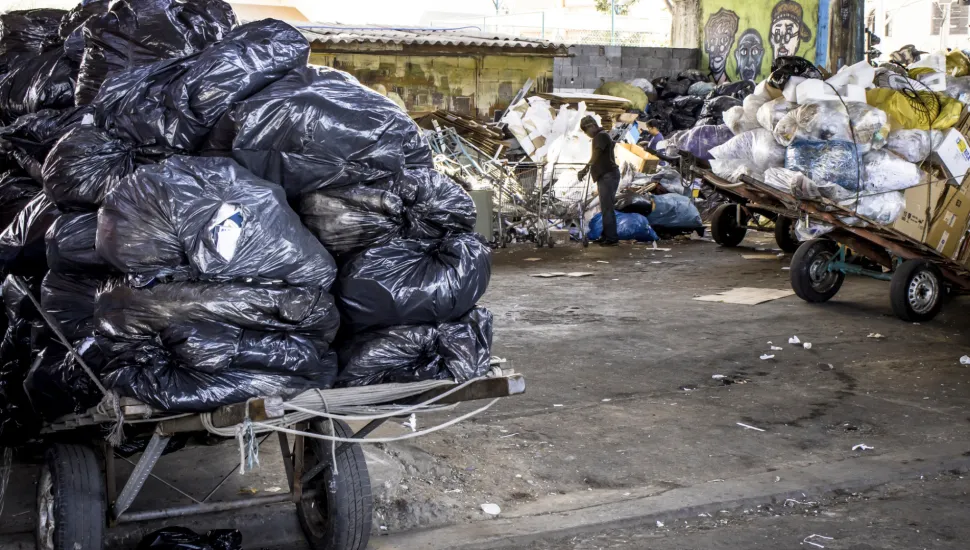 Fotografia em enquadramento amplo com plano geral de um ponto de coleta e triagem de materiais recicláveis localizado sob uma estrutura urbana de concreto. Em primeiro plano, à esquerda, destaca-se uma carroça de madeira equipada com pneus automotivos, empilhada com dezenas de sacos de lixo pretos cheios e amarrados por cordas brancas. No piso de concreto áspero e desgastado, veem-se pequenos resíduos e papéis espalhados. No plano intermediário, à direita, encontra-se outra carroça de tração manual carregada com caixas de papelão, placas de isopor e sacos transparentes contendo recicláveis. Ao centro-fundo, um trabalhador em pé, vestindo roupas escuras, manuseia e organiza resíduos ao lado de carrinhos de compras e grandes pilhas de materiais acumulados. Ao fundo, sob a estrutura do viaduto com pilares pintados com arte urbana e grafite, avistam-se construções simples e galpões sob luz diurna natural.