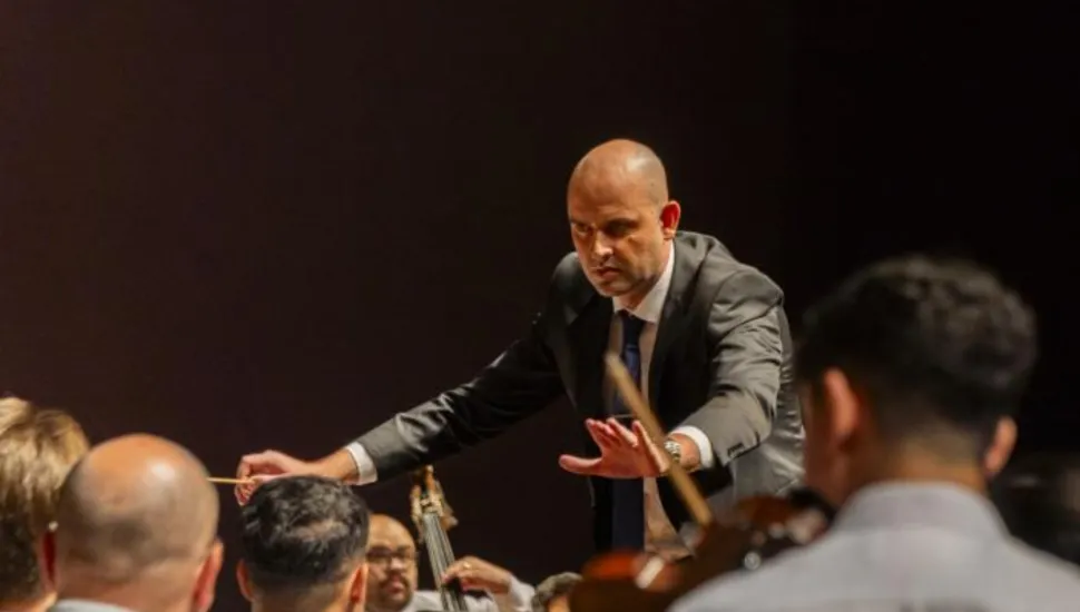 Fotografia de um regente de orquestra com os braços abertos coordenando os músicos, que são visto apenas de costas.