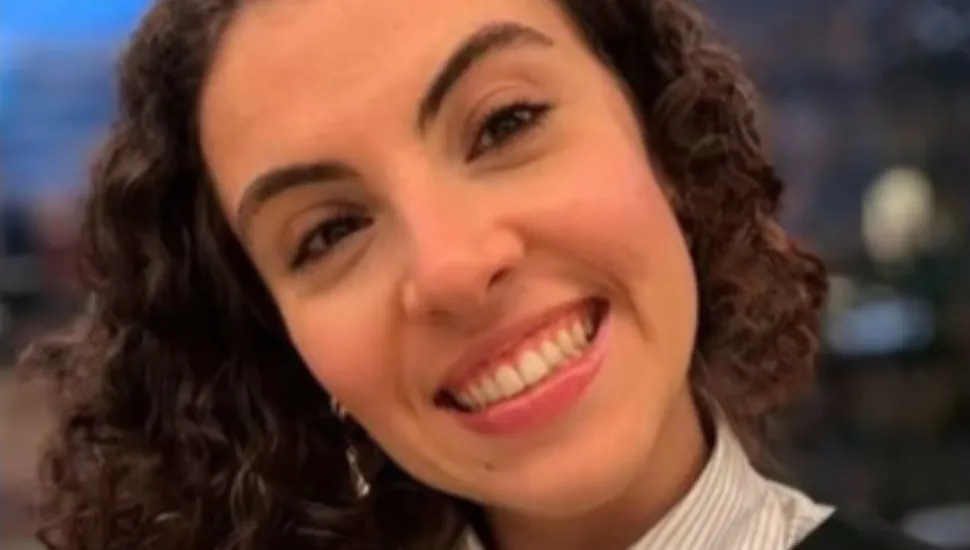 Fotografia em primeiro plano fechado (take aproximado) do rosto de uma mulher jovem sorrindo levemente inclinada para a direita. Ela tem a pele clara, olhos castanhos-escuros, sobrancelhas bem delineadas e cabelos cacheados volumosos em tom castanho-escuro que emolduram o rosto. Ela sorri abertamente para a câmera, exibindo os dentes superiores e usando batom ou brilho labial rosa-claro. Na parte inferior do enquadramento, observa-se a gola de uma camisa social listrada de tom claro sob uma sobreposição escura. O fundo encontra-se totalmente desfocado por conta da profundidade de campo rasa, com pontos suaves de iluminação em tom azulado e dourado no ambiente interno.