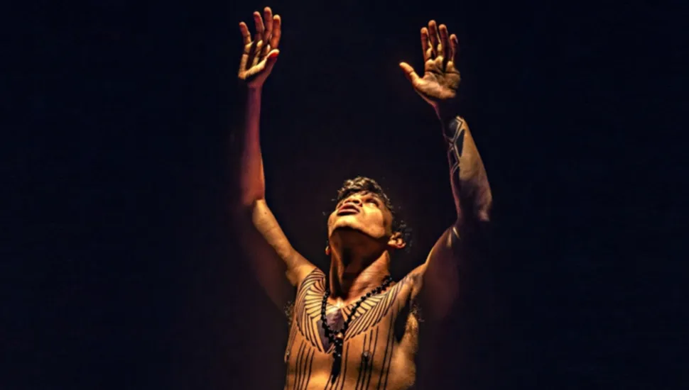 Fotografia dramática em plano médio de um homem indígena em cena artística ou performática sobre fundo escuro. Em primeiro plano, o homem é visto com o torso nu, exibindo pinturas corporais tradicionais com grafismos geométricos em linhas pretas no peito, abdômen e braço esquerdo, além de usar um colar de contas escuras no pescoço. Ele está com a cabeça inclinada para cima, olhar direcionado ao alto e os dois braços erguidos com as palmas das mãos voltadas para a frente em gesto expressivo. A iluminação é cênica e direcionada (foco de luz do tipo spot), destacando os contornos do corpo, os traços do rosto e as pinturas corporais contra o fundo preto e completamente escuro