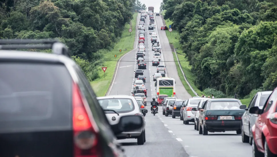 Fotografia em enquadramento amplo com plano geral do tráfego congestionado em um longo aclive de uma rodovia cercada por mata verde densa. Em primeiro plano, no canto esquerdo, observa-se em ângulo próximo e levemente desfocado a traseira de um SUV escuro com a lanterna vermelha ligada. Ao centro e à direita, alinham-se diversos carros de passeio em fila lenta, incluindo um sedan verde-escuro e um veículo vermelho. No plano intermediário, a rodovia de múltiplas faixas estende-se em uma longa subida recheada de veículos emparelhados. Um ônibus rodoviário branco e verde e um motociclista trafegam entre os carros ao longo do fluxo. À esquerda da pista, vê-se uma placa triangular vermelha de sinalização "Dê a Preferência". Ao fundo, o aclive da estrada perde-se no horizonte sob iluminação diurna e céu claro encoberto, margeado por encostas e floresta de vegetação nativa.