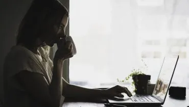 Imagem mostra uma mulher na frente de um notebook, tomando um café em frente a uma janela.