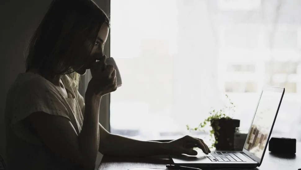 Imagem mostra uma mulher na frente de um notebook, tomando um café em frente a uma janela.