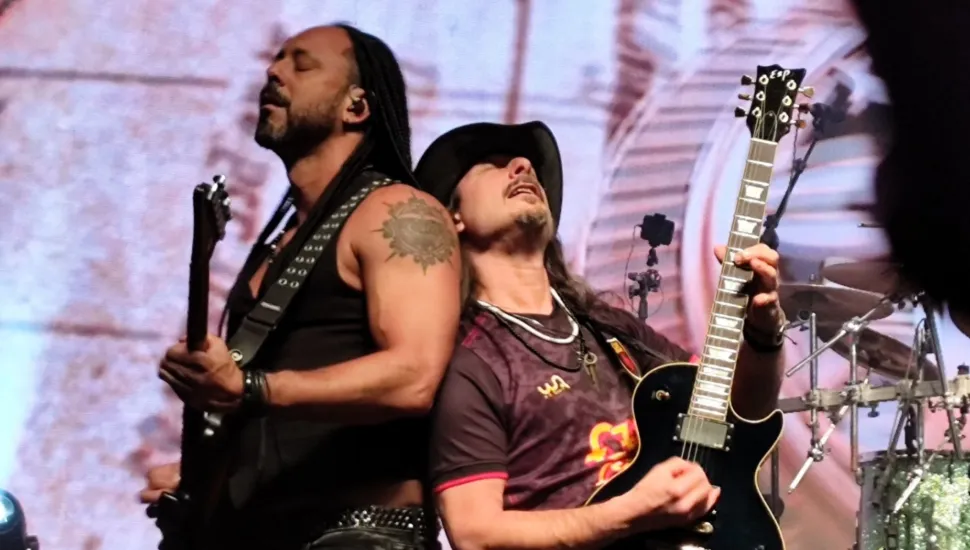 Fotografia em plano médio de dois músicos tocando guitarra costas com costas durante um show ao vivo em um palco. Em primeiro plano, à esquerda, um homem de pele morena com dreadlocks longos nos cabelos, barba e uma tatuagem circular no ombro esquerdo veste regata preta com tachas metálicas na alça. Ele está de perfil com a cabeça inclinada para trás e olhos fechados enquanto segura sua guitarra. Encostado a ele, à direita, outro homem de chapéu escuro, cavanhaque e cabelos longos veste camiseta estampada em tons de roxo e vermelho e segura uma guitarra elétrica preta da marca ESP com o braço elevado. No plano intermediário, à direita, avistam-se pratos e ferragens de uma bateria. Ao fundo, um painel cenográfico com texturas em tons claros de bege e iluminação cênica completa o cenário sob luzes do espetáculo.