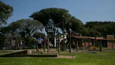 Fotografia em enquadramento amplo com plano geral de uma academia ao ar livre em uma praça ou parque público ensolarado. Em primeiro plano, estende-se uma área de gramado verde bem cuidado com uma caixa de areia delimitada por blocos de concreto. No centro, um homem de costas vestindo roupa esportiva azul e preta caminha em direção aos equipamentos de ginástica. No plano intermediário, diversas pessoas praticam atividades físicas em equipamentos de calistenia de madeira e estruturas metálicas para musculação e alongamento. À esquerda, praticantes realizam exercícios de alongamento, enquanto à direita frequentadores utilizam aparelhos da academia da terceira idade. Poste de iluminação pública e bicicletas estacionadas completam a cena. Ao fundo, sob céu azul sem nuvens, observam-se árvores de copas altas e frondosas, incluindo araucárias, além da cobertura de alvenaria e telha cerâmica das instalações do parque e uma encosta arborizada.