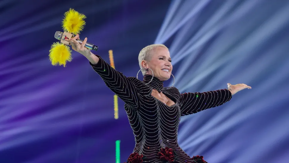 Fotografia em plano médio da apresentadora e cantora Xuxa Meneghel no centro de um palco durante uma apresentação ou show ao vivo. Em primeiro plano, enquadrada da cintura para cima, a artista de pele clara e cabelos loiros curtíssimos sorri com os braços abertos em tom de celebração. Ela veste um figurino elegante e ajustado em tom escuro (vinho ou preto) decorado com linhas onduladas reluzentes de strass, ombreiras marcadas, gola alta e detalhes de flores aveludadas vermelhas na altura da cintura. Ela usa grandes brincos de argola prateados e segura com a mão direita um microfone decorado com plumas amarelas nas extremidades. Ao fundo, a iluminação cênica exibe feixes de luz prateados e azulados em diagonal sobre um fundo arroxeado e luzes verticais coloridas dispostas no cenário.