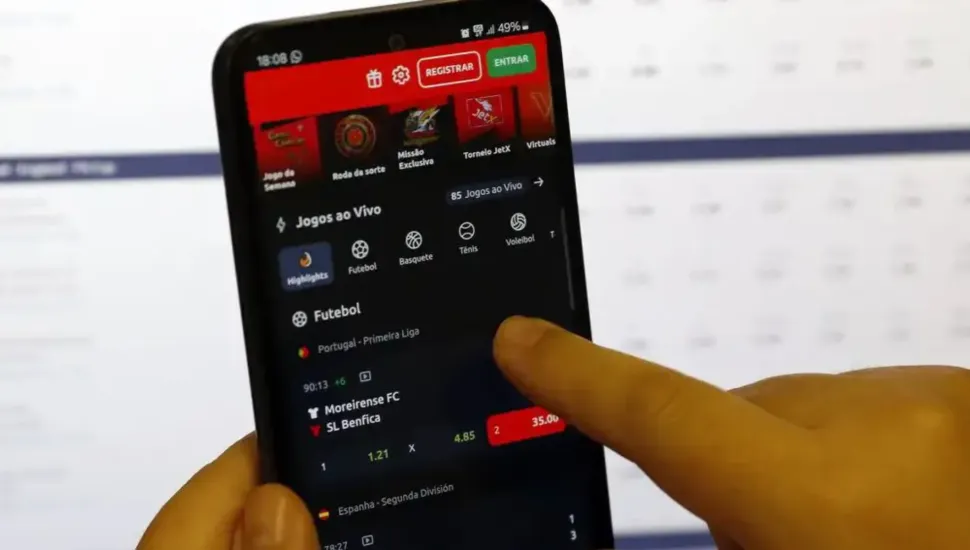 Fotografia em primeiro plano fechado de um smartphone sendo manuseado por uma pessoa enquanto exibe um aplicativo de apostas esportivas na tela. Em primeiro plano, à direita e abaixo, veem-se os dedos de uma mão humana segurando o dispositivo e tocando a tela sensível ao toque, apontando para as opções de apostas de uma partida de futebol. Na tela do celular, com interface em tema escuro e detalhes em vermelho e verde no topo, destacam-se seções como "Jogos ao Vivo" e o menu de esportes com ícones para Futebol, Basquete, Tênis e Voleibol. Abaixo, aparece a listagem de um jogo da "Portugal - Primeira Liga" entre Moreirense FC e SL Benfica, exibindo as cotações (odds) das apostas. Ao fundo, desfocado, observa-se o monitor de um computador ou tela clara com tabelas e dados numéricos sob iluminação artificial.