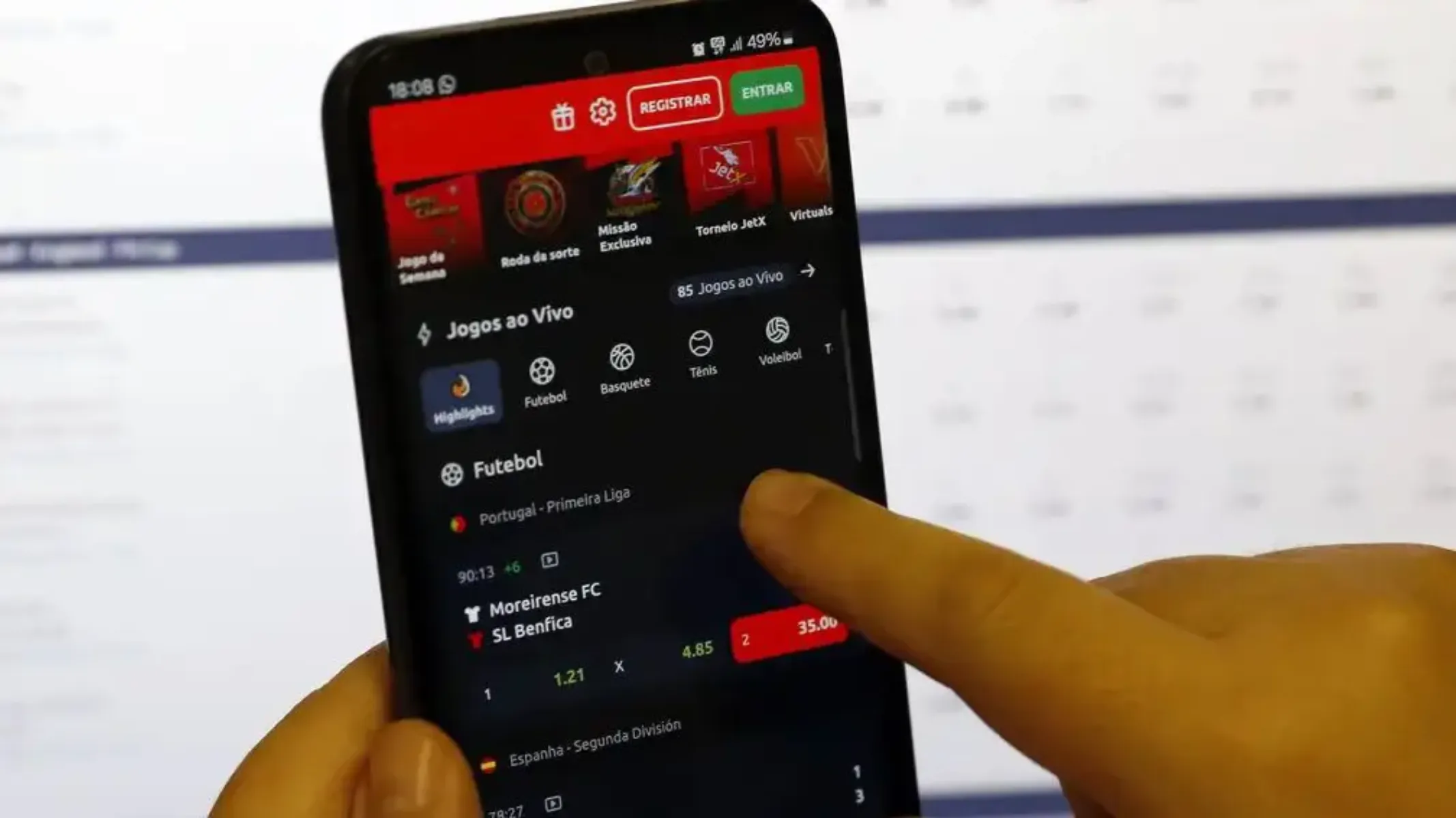Fotografia em primeiro plano fechado de um smartphone sendo manuseado por uma pessoa enquanto exibe um aplicativo de apostas esportivas na tela. Em primeiro plano, à direita e abaixo, veem-se os dedos de uma mão humana segurando o dispositivo e tocando a tela sensível ao toque, apontando para as opções de apostas de uma partida de futebol. Na tela do celular, com interface em tema escuro e detalhes em vermelho e verde no topo, destacam-se seções como "Jogos ao Vivo" e o menu de esportes com ícones para Futebol, Basquete, Tênis e Voleibol. Abaixo, aparece a listagem de um jogo da "Portugal - Primeira Liga" entre Moreirense FC e SL Benfica, exibindo as cotações (odds) das apostas. Ao fundo, desfocado, observa-se o monitor de um computador ou tela clara com tabelas e dados numéricos sob iluminação artificial.