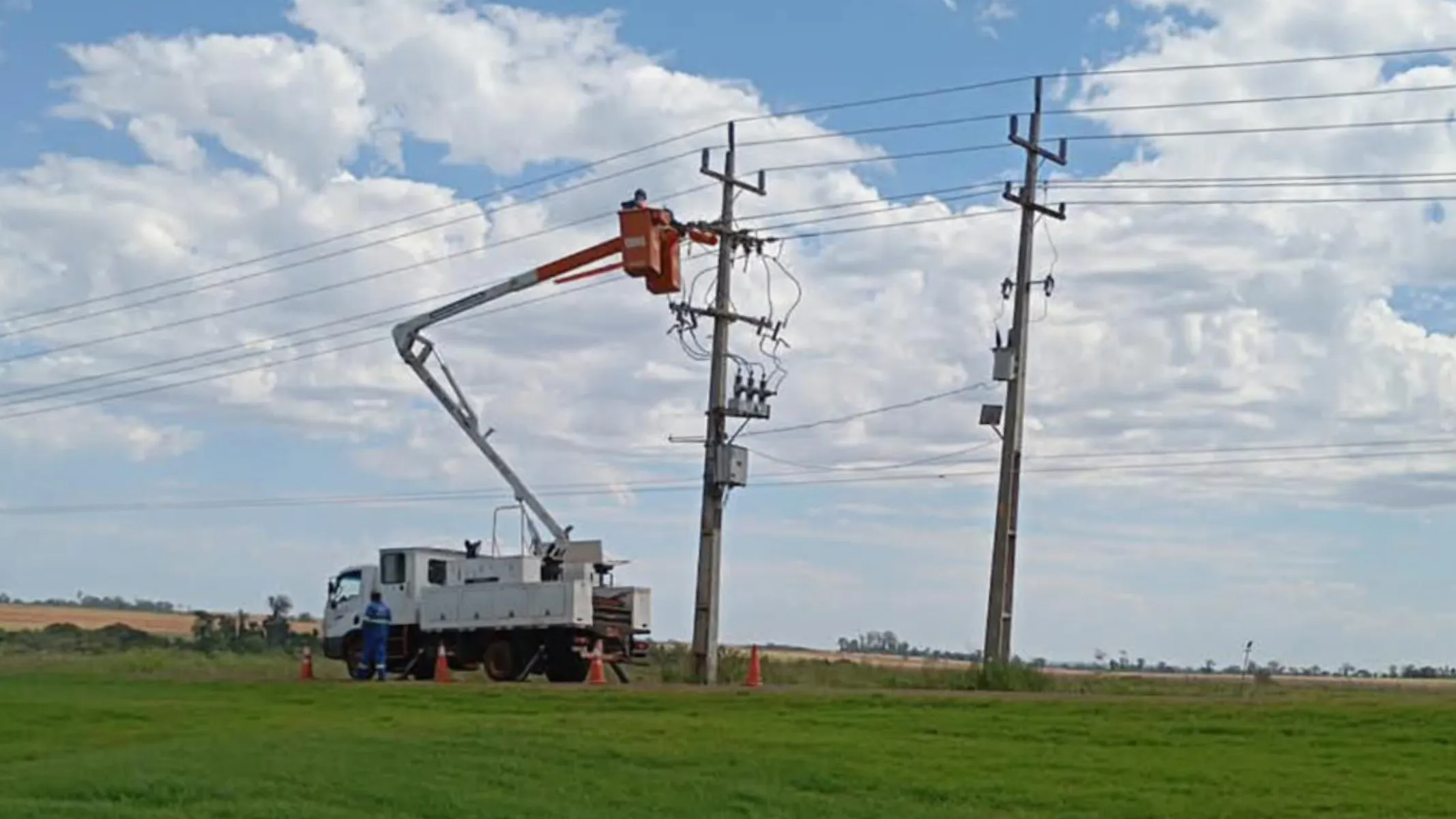 Fotografia em enquadramento amplo com plano geral de um serviço de manutenção e reparo na rede de energia elétrica realizado em uma área rural sob luz diurna natural. Em primeiro plano, estende-se uma ampla faixa de gramado verde em relevo plano. No plano intermediário, um caminhão utilitário de cor branca com caçamba de serviço está estacionado ao lado de uma estrada rural de terra. Cones de sinalização laranjas estão posicionados ao redor do veículo, e um eletricista de uniforme azul permanece em pé junto à lateral. O braço articulado telescópico do caminhão está elevado, sustentando um cesto aéreo Laranjado no qual um eletricista trabalha diretamente na fiação e nos equipamentos do topo de um poste de concreto da rede elétrica. Um segundo poste de concreto com transformador e fiação paralela situa-se à direita. Ao fundo, a paisagem descampada exibe vegetação Rasteira e campos agrícolas sob um céu azul coberto por nuvens brancas e volumosas.