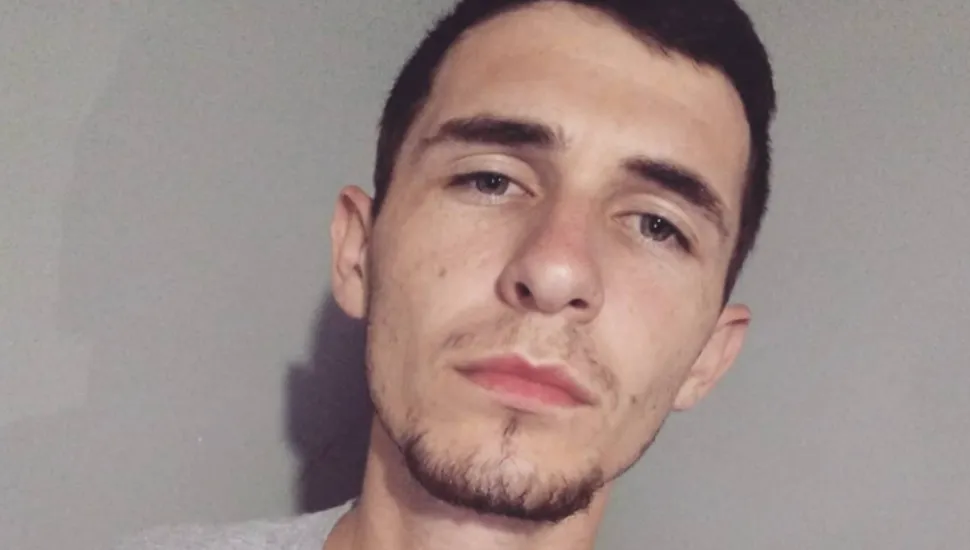 Fotografia em primeiro plano fechado do tipo autorretrato (selfie) de um jovem homem adulto, enquadrado do pescoço para cima, olhando diretamente para a câmera. Em primeiro plano, o jovem possui pele clara, rosto de formato oval, cabelos castanho-escuros curtos e sobrancelhas marcadas. Ele apresenta olhos claros, barba rala por fazer desenhando o maxilar e o queixo, além de lábios entreabertos em uma expressão neutra e séria. Na parte inferior esquerda do enquadramento, observa-se a gola de uma camiseta cinza-clara. Ao fundo, sob iluminação frontal uniforme que projeta uma sombra suave à esquerda da imagem, destaca-se uma parede lisa e plana em tom cinza-neutro.