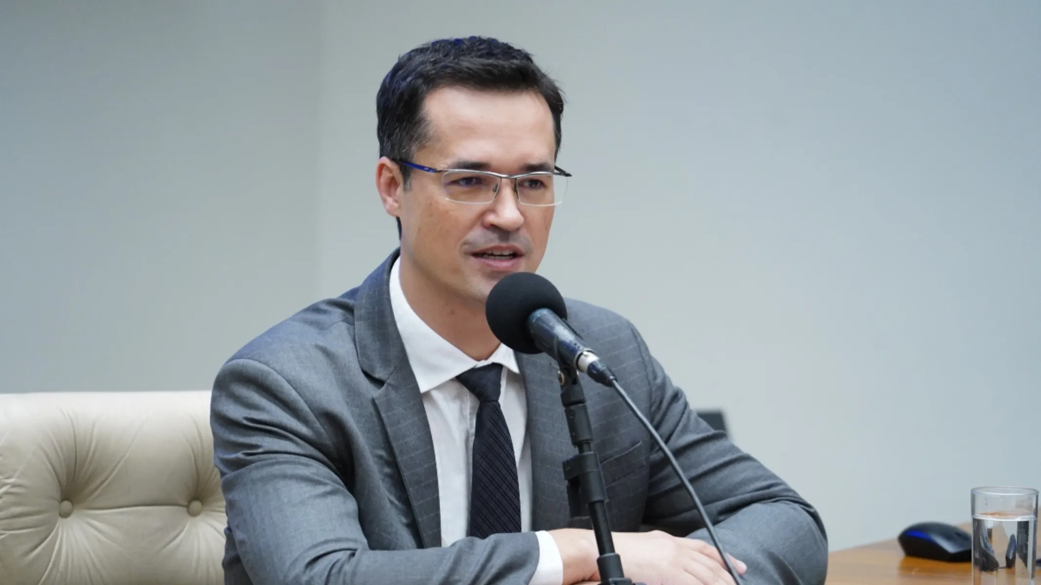 Fotografia em plano médio do ex-procurador da República Deltan Dallagnol discursando em uma mesa de conferência ou reunião institucional. Em primeiro plano, enquadrado do peito para cima, um homem de pele clara, cabelos pretos curtos penteados para o lado e óculos de armação fina sem aro inferior fala diante de um microfone pedestal de espuma preta posicionando no centro da mesa. Ele veste um terno cinza-médio com padronagem risca-de-giz discreta, camisa social branca e gravata preta texturizada. Ele apoia os braços sobre a mesa de madeira clara, onde se observam um copo com água e um mouse de computador no canto inferior direito. No plano intermediário, à esquerda, sobressai o encosto de uma poltrona executiva estofada em couro de cor bege claro com acabamento capitonê. Ao fundo, sob iluminação uniforme de ambiente corporativo, destaca-se uma parede lisa de tom bege claro e neutro.