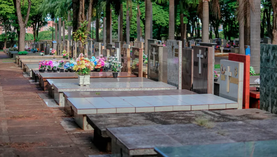 Fotografia em enquadramento amplo com perspectiva em diagonal do corredor de um cemitério sob luz natural diurna. Em primeiro plano, à esquerda, estende-se uma passagem pavimentada em lajotões de concreto em tom avermelhado. À direita, sucedem-se sepulturas horizontais revestidas em lajotas de cerâmica e granito claro, com lápides verticais ornamentadas com crucifixos e símbolos religiosos. No plano intermediário, sobre as lajes dos túmulos, observam-se diversos vasos ornamentados com flores artificiais coloridas em tons de rosa, amarelo, azul e branco. Ao fundo, fileiras paralelas de túmulos continuam sob a sombra de altas palmeiras e árvores de copas frondosas e verdes que arborizam o local.