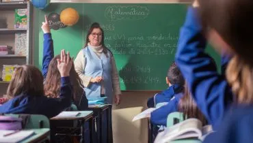 A imagem mostra uma professora em sala de aula ao fundo e alunos com as mãos levantadas.