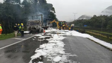 Fotografia em enquadramento amplo com plano geral do atendimento a um acidente com incêndio de caminhão em uma rodovia sob céu nublado e chuva fina. Em primeiro plano, a pista de asfalto molhado estende-se em perspectiva diagonal, coberta por densa camada de espuma branca de combate a incêndio espalhada pela pista e pelas margens. No plano intermediário, à esquerda, a carcaça totalmente queimada e destruída de um caminhão de carga emite fumaça. Ao lado do veículo, agentes do Corpo de Bombeiros com capacetes amarelos e trajes de proteção e um agente da Polícia Rodoviária Federal (PRF) com jaqueta escura operam no local. Logo atrás do caminhão, uma retroescavadeira amarela atua na remoção de destroços da via. À direita, muretas de concreto delimitam as faixas da rodovia. Ao fundo, a paisagem é cercada por mata nativa e morros cobertos por vegetação densa sob neblina e céu cinzento.