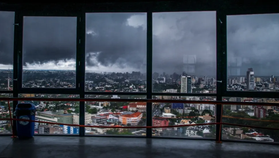 Fotografia em enquadramento amplo com plano geral panorâmico da vista urbana de uma cidade observada através de grandes janelas de vidro em um ponto de observação elevado, sob tempo severo e tempestuoso. Em primeiro plano, na parte inferior e interna do ambiente, estende-se o piso escuro de cimento e uma barra de proteção/guarda-corpo na cor vermelha que corta horizontalmente a extensão das janelas. À esquerda, junto à estrutura metálica da janela, há uma lixeira cilíndrica de cor azul. No plano intermediário, dividida por caixilhos retangulares pretos das janelas de vidro, sobressai a vista aérea do espaço urbano, composta por diversos edifícios, casas, ruas e vegetação densa intercalada entre as construções. Ao fundo, o horizonte e o céu são cobertos por densas e escuras nuvens de tempestade em tons de cinza-chumbo, sinalizando chuva forte sobre a região central e periférica da cidade.
