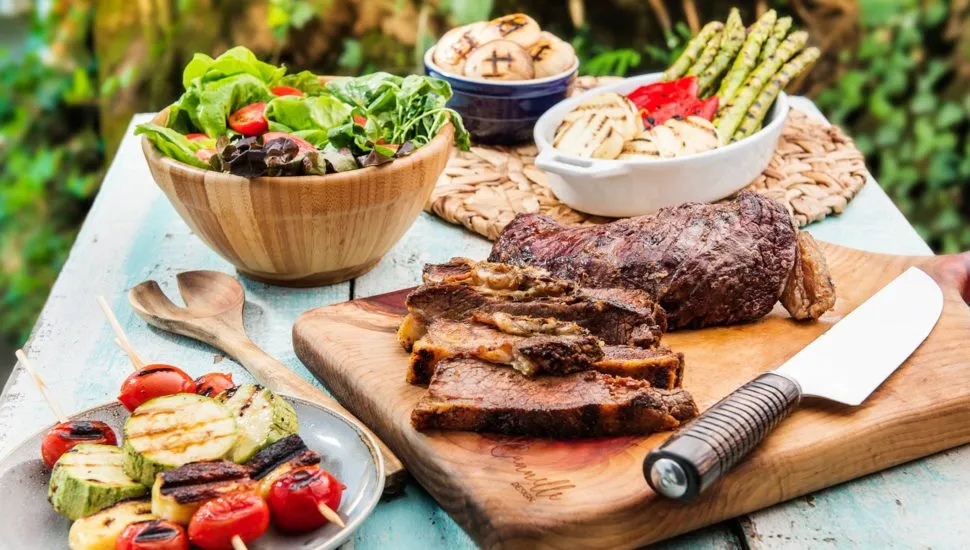 Imagem mostra um churrasco completo, com carne, salada e acompanhamentos em uma mesa.