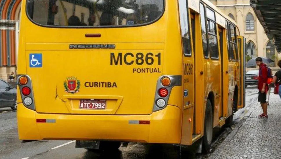Fotografia em enquadramento médio com perspectiva em diagonal traseira de um ônibus do transporte coletivo urbano na cor amarela, estacionado em um ponto de embarque. Em primeiro plano, ocupa o centro da imagem a traseira do veículo amarelo, onde se destacam o código "MC861", a palavra "PONTUAL", o brasão oficial e a inscrição "CURITIBA", além do símbolo internacional de acessibilidade em fundo azul e a placa de licenciamento "ATC-7992". No plano intermediário, à direita, a lateral do ônibus apresenta suas portas abertas em uma estação de embarque sobre piso de paralelepípedos molhado pela chuva. Próximo às portas, um jovem segura um guarda-chuva e veste casaco vermelho com mochila nas costas. Ao fundo, sob céu nublado, vislumbra-se a fachada de um prédio histórico de arquitetura clássica e um veículo cinza estacionado na rua.
