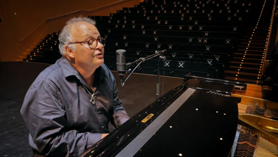 Fotografia em plano médio do cantor, compositor e pianista Guilherme Arantes tocando piano em um palco de teatro iluminado. Em primeiro plano, enquadrado do peito para cima e posicionado de perfil no canto esquerdo, sobressai Guilherme Arantes, um homem de pele clara, cabelos grisalhos curtos e encaracolados e óculos de armação redonda escuro. Ele veste uma camisa social cinza-escura com os primeiros botões abertos, revelando um colar com pingente em formato de nota musical. Ele canta de boca aberta direcionado a um microfone de estúdio prateado montado em pedestal articulado. Suas mãos estão sobre o teclado de um piano de cauda preto brilhante que ocupa a metade inferior direita da imagem. No plano intermediário e ao fundo, vê-se a vasta plateia vazia de um teatro de grande porte disposta em formato de anfiteatro ascendente. As poltronas pretas apresentam marcações em formato de "X" nos encostos, sob iluminação cênica suave e pontual nos degraus de acesso às galerias.