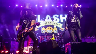 Fotografia em enquadramento médio com contra-plano de uma apresentação musical de rock em palco com iluminação cênica de tom roxo e violeta. Em primeiro plano, à esquerda, destaca-se o guitarrista Slash, de pele morena, cabelos pretos cacheados volumosos cobrindo parcialmente o rosto e vestindo sua tradicional cartola preta, óculos escuros, camiseta preta estampada e calça de couro preta. Ele está inclinado para a frente enquanto toca uma guitarra elétrica Les Paul escura. À direita, no mesmo plano, o vocalista Myles Kennedy canta em um microfone fixado em um pedestal inclinado. Ele veste jaqueta de couro escura, camiseta estampada e calça preta. No plano intermediário, entre os dois músicos e ao fundo, vê-se a bateria com um integrante ao fundo e o painel de fundo (backdrop) do show, exibindo a palavra "SLASH" em letras maiúsculas estilizadas com contorno azul e a inscrição "FEATURING MYLES KENNEDY AND THE CONSPIRATORS". Ao fundo, potentes refletores de luz de palco no topo e na base emitem holofotes luminosos sob atmosfera envolta em fumaça cênica roxa.