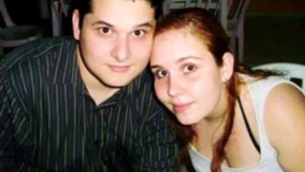Fotografia em primeiro plano do tipo retrato aproximado de um casal jovem posando lado a lado com as cabeças encostadas. Em primeiro plano, à esquerda, destaca-se um homem de pele clara e cabelos escuros curtos, vestindo uma camisa social de mangas compridas preta com finas listras verticais em tom claro/esverdeado. À direita, enquadrada do peito para cima e com a cabeça apoiada suavemente na dele, está uma mulher de pele clara, olhos castanhos e cabelos castanho-claros amarrados para trás, vestindo uma regata branca de alças largas sobre top preto de alças finas. Ambos olham diretamente para a câmera com expressões neutras e leves sorrisos. No plano intermediário e ao fundo, ligeiramente desfocados, veem-se cadeiras e mesas de plástico branco em um ambiente social sob iluminação indireta/noturna.
