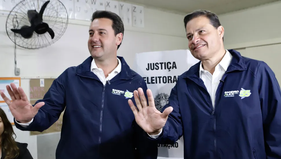 Fotografia em plano médio do governador do Paraná, Ratinho Junior, ao lado do político Sandro Alex em uma seção eleitoral durante votação. Em primeiro plano, no centro da imagem, posicionam-se dois homens sorridentes enquadrados da cintura para cima, vestindo jaquetas idênticas em tom azul-marinho sobre camisas sociais brancas. No peito esquerdo das jaquetas há uma estampa com a inscrição "SANDRO ALEX" acompanhada do número "55" em amarelo e verde. À esquerda, Ratinho Junior, homem de pele clara e cabelos castanho-escuros curtos penteados para trás, acena levantando a mão direita com os dedos abertos em direção à câmera. À direita, Sandro Alex, homem de pele clara e cabelos castanho-escuros curtos, também faz um gesto de aceno levantando a mão direita espalmada para a frente. No plano intermediário, situado entre os dois homens, destaca-se uma cabina de votação de papelão branco com a inscrição "JUSTIÇA ELEITORAL" em letras maiúsculas pretas acima do Brasão da República. No canto superior esquerdo da parede, observa-se um ventilador de parede preto e cartazes escolares fixados. Ao fundo, a sala de aula adaptada como seção eleitoral apresenta iluminação artificial fria e uniforme.