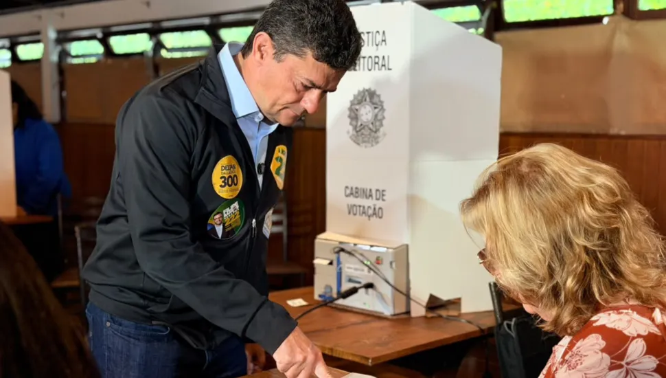 Fotografia em plano médio do senador Sergio Moro em uma seção eleitoral no momento de identificação antes da votação. Em primeiro plano, no lado esquerdo da imagem, destaca-se Sergio Moro, um homem de pele clara e cabelos castanho-escuros curtos, visto de perfil inclinado para a frente e olhando para baixo. Ele veste uma jaqueta esportiva preta com zíper sobre camisa azul-clara e calça jeans. No peito de sua jaqueta há diversos adesivos de campanha eleitoral afixados. Com a mão direita estendida para baixo, ele aponta ou assina a lista de votantes sobre a mesa. No canto inferior direito, em primeiro plano, vê-se de costas a cabeça e os ombros de uma mesária de cabelos loiros ondulados usando blusa estampada floral. No plano intermediário, sobre uma mesa de madeira ao centro, posiciona-se a urna eletrônica conectada ao terminal do eleitor, parcialmente coberta por uma cabina de votação de papelão branco com as inscrições "JUSTIÇA ELEITORAL", o Brasão da República e a expressão "CABINA DE VOTAÇÃO". Ao fundo, observa-se o interior do local de votação com janelas altas e iluminação natural suave.