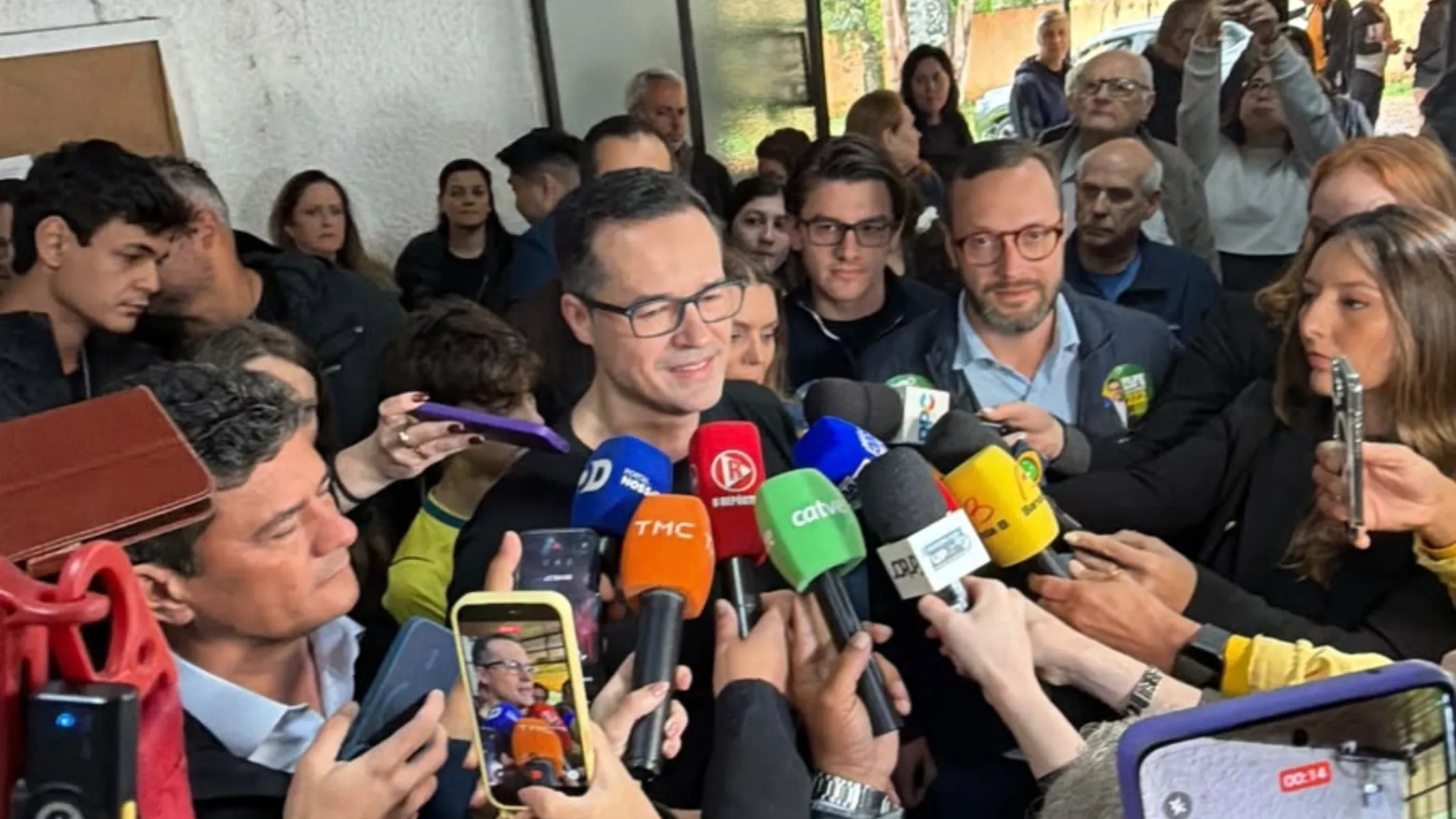 Fotografia em plano médio de uma entrevista coletiva de imprensa com grande aglomeração de jornalistas e políticos em ambiente interno. Em primeiro plano, no centro da imagem, destaca-se o ex-procurador e político Deltan Dallagnol, um homem de pele clara, cabelos castanho-escuros curtos e óculos de armação fina, sorrindo enquanto fala cercado por diversos microfones de veículos de imprensa (com canoplas coloridas em azul, laranja, verde e amarelo) e celulares apontados para gravá-lo. No canto inferior esquerdo, enquadrado de perfil e também cercado por repórteres, está o senador Sergio Moro, homem de pele clara e cabelos escuros. No canto inferior direito, uma repórter segura um smartphone registrando a entrevista. No plano intermediário, atrás de Deltan Dallagnol, observam-se assessores e outros políticos acompanhando a coletiva, incluindo o deputado estadual Denian Couto de óculos e camisa clara. Ao fundo, uma multidão de apoiadores, eleitores e populares aglomera-se no espaço sob iluminação natural suave vinda de uma abertura ao fundo.